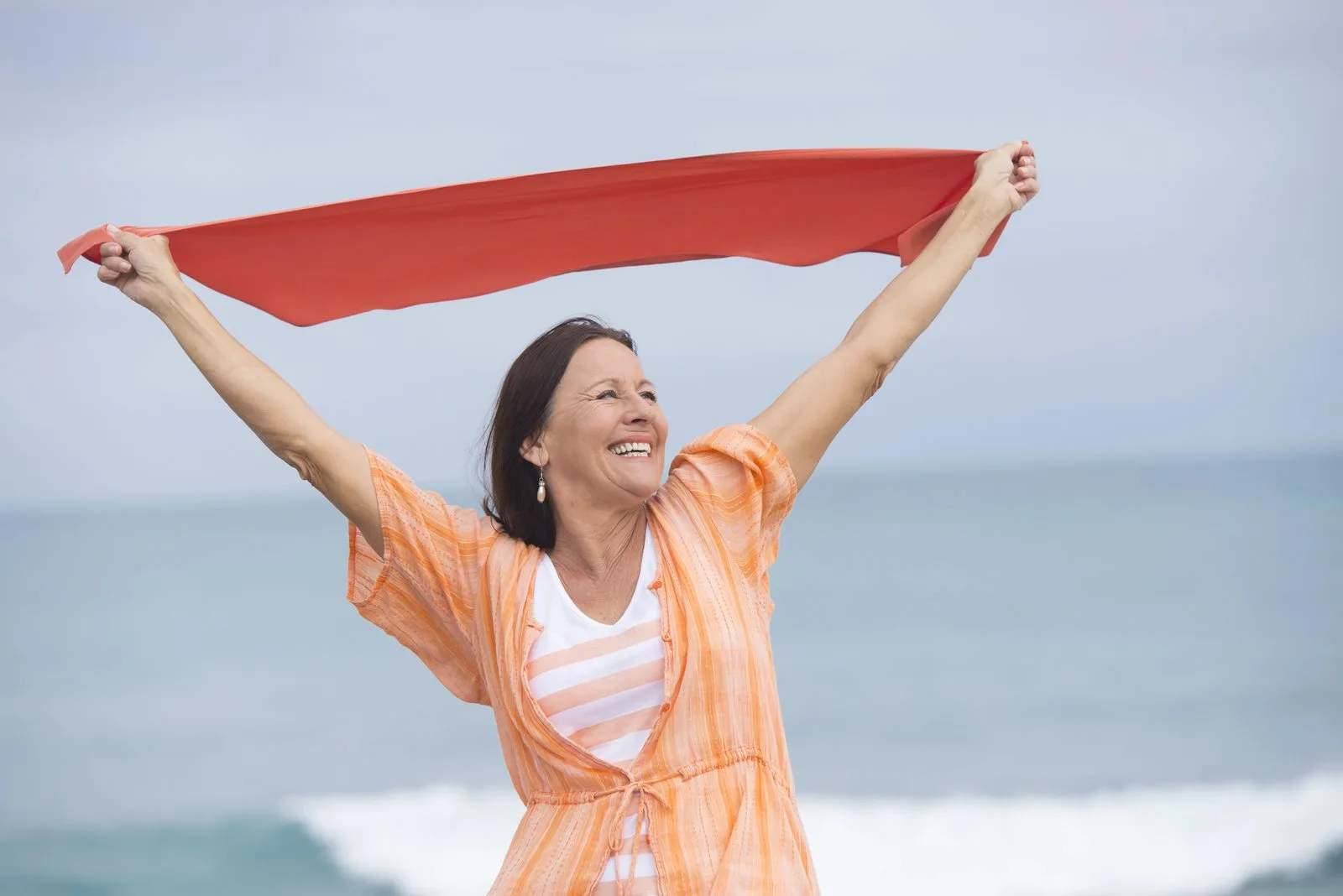 Lachende Frau am Meer hält ein rotes Tuch im Wind – Fasten in den Wechseljahren