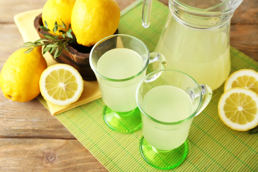 Zwei Gläser und eine Karaffe mit Zitronenwasser, daneben frische Zitronen auf einer grünen Bastmatte