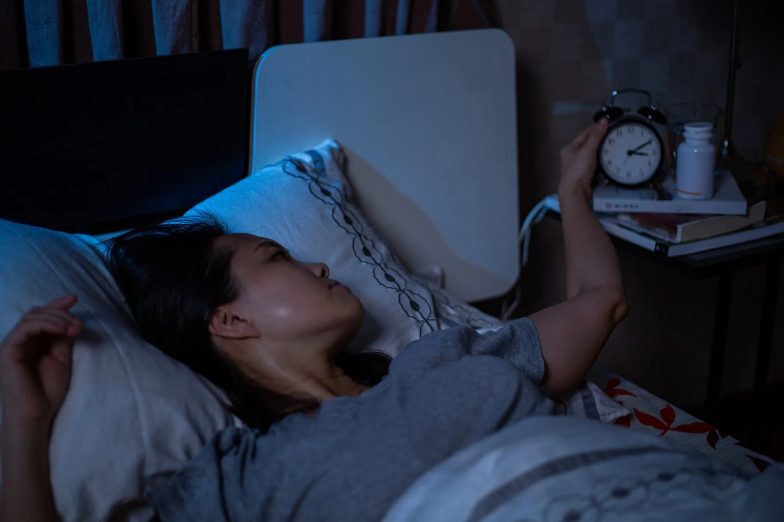 Frau liegt nachts wach im Bett und schaut auf den Wecker – Schlafstörungen und Fasten